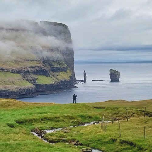 Ein Landschaftsbild von den Färöer Inseln. Mittig steht eine Person am Rande eines Feldes an einer Klippe, die an einem Fjord liegt. Auf der anderen Fjordseite sieht man schroffe Klippen und die Felsnadeln von Risin og Kellingin. 