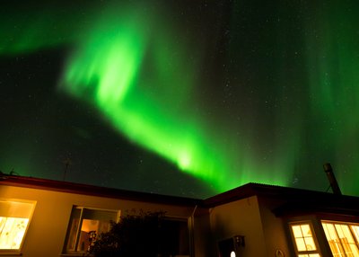 Grellgrün tanzt ein Polarlicht über einem beleuchteten Haus. Die Sterne scheinen am Nachthimmel