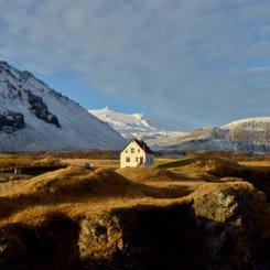 Im Abendlicht sieht man eine Winterlandschaft, inmitten dieser ein Haus steht. Im Hintergrund leuchtet die Spitze des Gletschers Snæfellsjökull