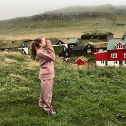 Ein Kind mit langen Haaren und in einem rosa Anzug steht auf einer Wiese vor einem roten Haus und schaut durch ein Fernglas. 