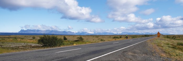 Panoramabild einer Landschaft in Island. Im Fordergrund sieht man eine Straße, an der auf der gegenüberleigenden Seite eine Radfahrerin mit ihrem Rad steht.