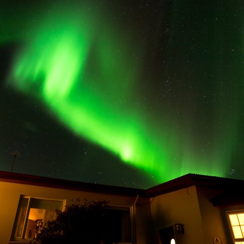 Grellgrün tanzt ein Polarlicht über einem beleuchteten Haus. Die Sterne scheinen am Nachthimmel