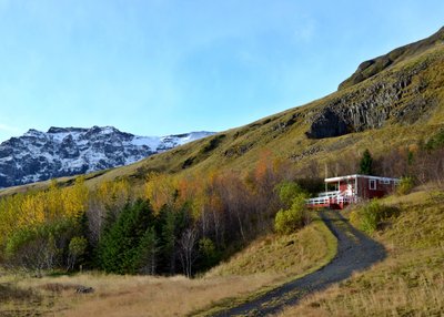 Ein rotes Holzhaus steht an einem Hang. Vorgelagert sind herbstbunte Bäume. Im Hintergrund sieht man einen schneebedeckten Berg.