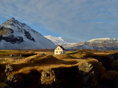 Im Abendlicht sieht man eine Winterlandschaft, inmitten dieser ein Haus steht. Im Hintergrund leuchtet die Spitze des Gletschers Snæfellsjökull