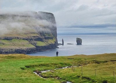 Ein Landschaftsbild von den Färöer Inseln. Mittig steht eine Person am Rande eines Feldes an einer Klippe, die an einem Fjord liegt. Auf der anderen Fjordseite sieht man schroffe Klippen und die Felsnadeln von Risin og Kellingin. 