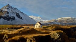 Im Abendlicht sieht man eine Winterlandschaft, inmitten dieser ein Haus steht. Im Hintergrund leuchtet die Spitze des Gletschers Snæfellsjökull