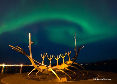 Über der Statue Sun Voyager in Reykjavik kringelt sich ein grünes Polarlicht. Das Zeichen des Großen Wagen ist auch zu erkennen.