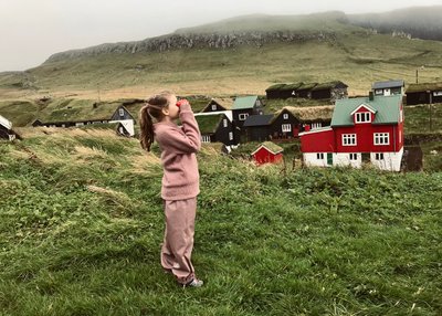 Ein Kind mit langen Haaren und in einem rosa Anzug steht auf einer Wiese vor einem roten Haus und schaut durch ein Fernglas. 