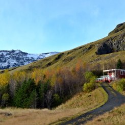 Ein rotes Holzhaus steht an einem Hang. Vorgelagert sind herbstbunte Bäume. Im Hintergrund sieht man einen schneebedeckten Berg.