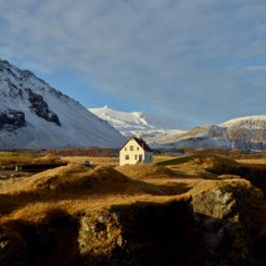 Im Abendlicht sieht man eine Winterlandschaft, inmitten dieser ein Haus steht. Im Hintergrund leuchtet die Spitze des Gletschers Snæfellsjökull