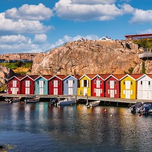 Schwedische bunte Holzhäuser stehen an einem kleinen Hafen. Im Wasser befinden sich ein paar kleine am Steg angelegte Boote.