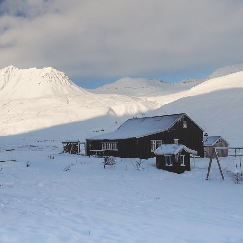Das schwarze Haus der Unterkunft im Svarfadardalur steht inmitten einer weißen Winterlandschaft. Im Hintergrund leuchtet ein Berg weiß.