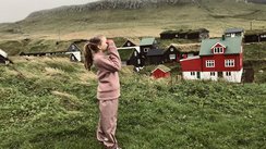 Ein Kind mit langen Haaren und in einem rosa Anzug steht auf einer Wiese vor einem roten Haus und schaut durch ein Fernglas. 